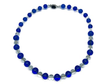 Op de afbeelding: Een ketting met afwisselend blauwe en heldere glazen kralen. De kralen zijn rond en geregen met een zilverkleurige sluiting. De ketting wordt gepresenteerd tegen een witte achtergrond.
