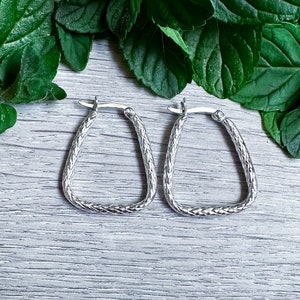 May include: A pair of silver hoop earrings with a textured, woven design. The earrings are in the shape of a triangle.