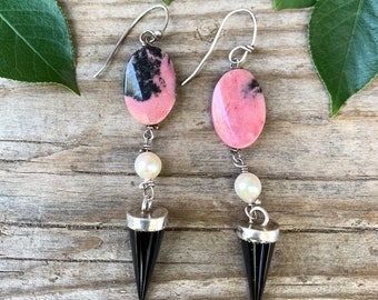May include: A pair of dangle earrings featuring pink and black oval-shaped stones, white pearl accents, and black pointed tips.