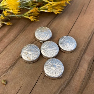 May include: Five silver metal round buttons with a daisy design. The buttons are on a wooden surface.