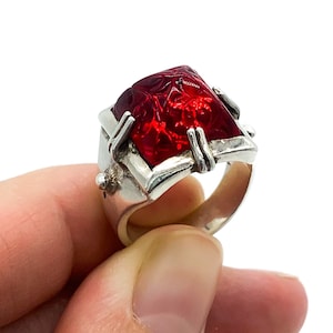 May include: A silver ring featuring a large, red, faceted gemstone. The gemstone has a carved design and is set in a silver band with intricate details.