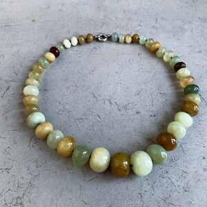 Puede incluir: Un collar hecho de cuentas de jade verde, marrón y blanco. Las cuentas son redondas y tienen un acabado liso y pulido.
