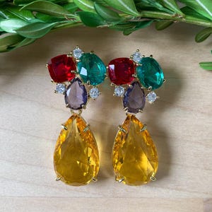 May include: A pair of gold-toned earrings featuring a cluster of colorful gemstones. The earrings have red, green, purple, and yellow teardrop-shaped stones. Small clear crystals accent the design. The earrings are set against a light wooden surface.