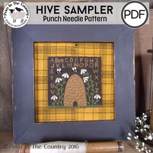 Op de afbeelding: Een ingelijst borduurwerk met een bijenkorf, bijen en madeliefjes. Het borduurwerk is op een gele en bruine geruite stof. De tekst "HIVE SAMPLER" en "Punch Needle Pattern" staat boven het borduurwerk. De tekst "Prim In The Country 2016" staat onder het borduurwerk.