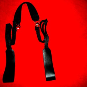 May include: A black leather strap with two metal buckles and a loop at the top. The strap is hanging against a red background.