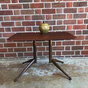May include: A mid-century modern table with a dark wood top and a metal base. The table has a unique tripod design with three legs that extend out from the center of the base. The table is sitting on a concrete floor in front of a brick wall.
