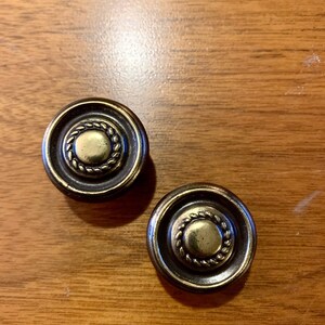 May include: Two round, brass cabinet knobs with a decorative, rope-like design around the edge. The knobs have a gold-colored center.