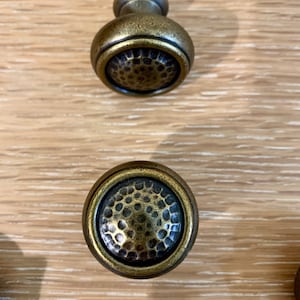 May include: Two antique brass cabinet knobs with a hammered finish. The knobs are round and have a raised edge.