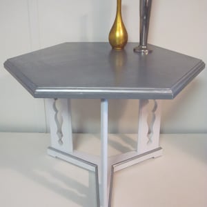 May include: A silver hexagonal table with a white base and decorative cutouts. The table has a silver top and white legs with a silver trim.