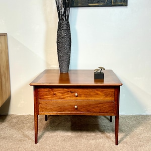 May include: A square wooden side table with a rich brown finish, featuring two drawers with small round brass-colored knobs. The table stands on four tapered legs and has a smooth, flat top surface. A tall, textured black vase and a small mushroom-shaped ornament sit on the table.