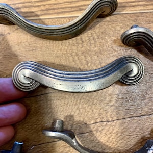 May include: Set of six antique silver cabinet drawer pulls with a decorative, wavy design. The pulls are made of metal and have a brushed finish.