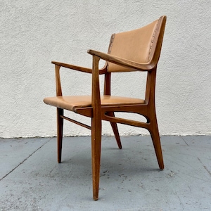 May include: A mid-century modern armchair with a light brown leather seat and back. The chair has a wooden frame with a unique design and angled legs.
