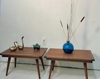 May include: Two mid-century modern wooden side tables with angled legs. One table has a blue glass vase with dried stems. The other table has brass swan figurines and a small teal ceramic dish.