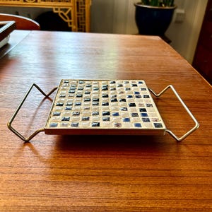 May include: A square ceramic tile coaster with a black and white checkered pattern. The coaster is on a metal frame with two handles.
