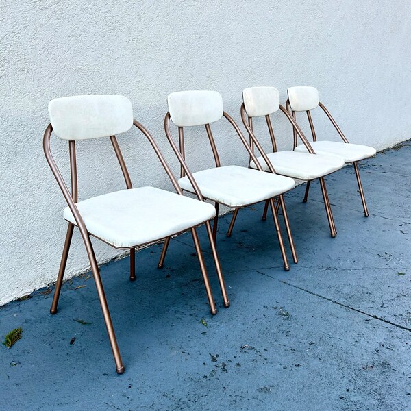 May include: Four white folding chairs with a rose gold metal frame. The chairs are lined up in a row on a concrete surface.
