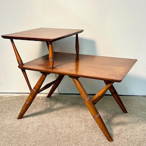 May include: A two-tiered, mid-century modern side table. The table is constructed of wood with a warm, medium-brown finish. The top tier is smaller than the lower one. The legs have a unique, angled design. The table measures approximately 30 inches long.