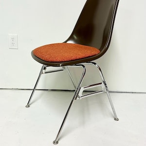 May include: A mid-century modern chair with a brown plastic shell and an orange fabric seat. The chair has a chrome-plated steel base with four legs. The design is sleek and minimalist, suitable for various interior styles.