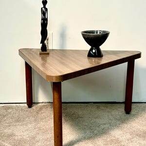May include: A mid-century modern triangular side table with a wood grain finish. It has three brown legs and a black sculpture of a woman. A black bowl with a speckled interior sits on the table.