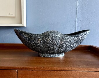 May include: A black and white speckled ceramic bowl with a unique, asymmetrical shape. The bowl is sitting on a wooden surface.