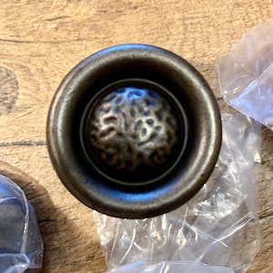 May include: A round, bronze-colored cabinet knob with a textured, circular center. The knob has a raised, beveled edge.