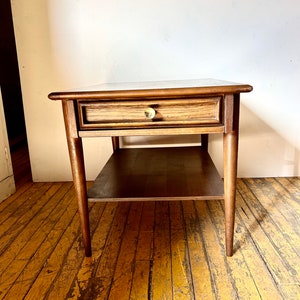 May include: A mid-century modern end table with a single drawer and a lower shelf. The table is made of wood with a dark brown finish. The drawer has a brass knob.