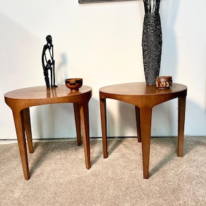 May include: Two wooden side tables with a light brown finish. Each table has a rounded top and four tapered legs. Decorative items, including a black sculpture and a small bowl, are displayed on the tables. A tall black vase is also visible.