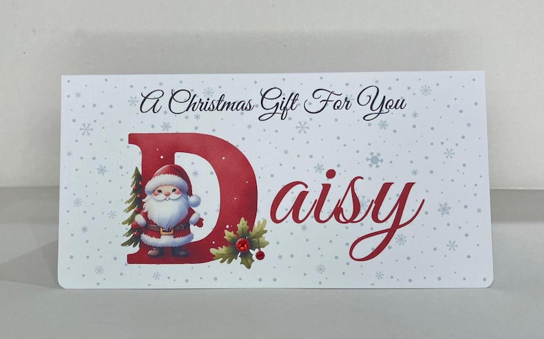 May include: A rectangular Christmas gift card with a festive design. The card features a red letter "D" with a Santa Claus illustration, the name "Daisy" in cursive, and the text "A Christmas Gift For You". Snowflakes are scattered across the white background.