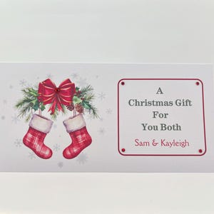 May include: A Christmas gift card featuring a festive design. The card displays two red plaid Christmas stockings hanging below a red bow and greenery. Text on the card reads "A Christmas Gift For You Both" and the names "Sam & Kayleigh".