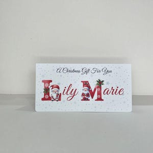 May include: A white rectangular gift card with the text "A Christmas Gift For You". The name "Lily Marie" is in red, with Santa Claus illustrations. The background has a snowflake pattern.