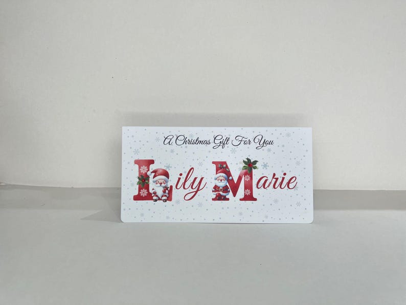 May include: A white rectangular gift card with the text "A Christmas Gift For You". The name "Lily Marie" is in red, with Santa Claus illustrations. The background has a snowflake pattern.