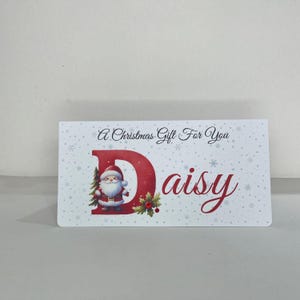 May include: A rectangular Christmas gift card with the text "A Christmas Gift For You" and the name "Daisy". The letter "D" is red and features a Santa Claus illustration. The card has a white background with snowflake accents.