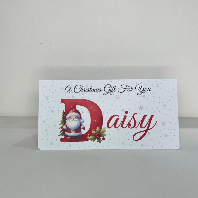May include: A rectangular Christmas gift card with the text "A Christmas Gift For You" and the name "Daisy". The letter "D" is red and features a Santa Claus illustration. The card has a white background with snowflake accents.