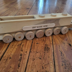 May include: A wooden toy train with a rectangular car and round wheels. The train car has a long, open top and a small wooden sign that reads "ANY NAME HERE". The train is made of light-colored wood and sits on a wooden surface.