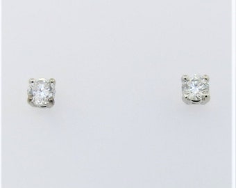 Puede incluir: Un par de pequeños pendientes de diamantes. Cada pendiente presenta un único diamante claro y brillante engastado en un metal de color plateado. Los pendientes se muestran sobre un fondo blanco liso.