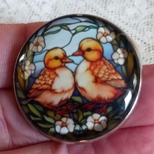 May include: A round, silver-rimmed pendant featuring a stained-glass style design. Two yellow and orange birds are depicted, surrounded by white flowers and green foliage against a blue background. The pendant is held in a hand.