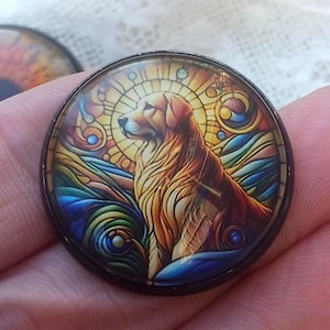 May include: A round, black-rimmed pendant featuring a Golden Retriever in a stained-glass style design. The dog is depicted in shades of gold, brown, and orange, set against a backdrop of blue, yellow, and green. The pendant is held in a hand.