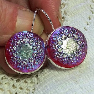 May include: A pair of silver earrings with a round, iridescent pink and purple glass design. The glass has a textured floral pattern.
