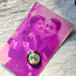 May include: A vintage postcard with a sepia-toned image of a couple, with a button featuring a cartoon image of a cherub holding an umbrella with hearts. The postcard is purple with the words "Bonne Pensee" in cursive.