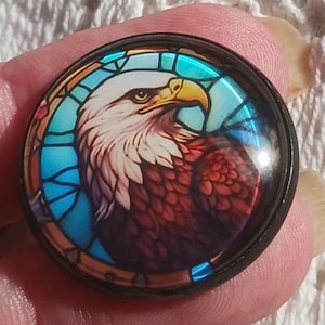 May include: Round, black-rimmed button with a stained-glass style image of a bald eagle. The eagle has a white head, yellow beak, and brown and red feathers, set against a blue background.