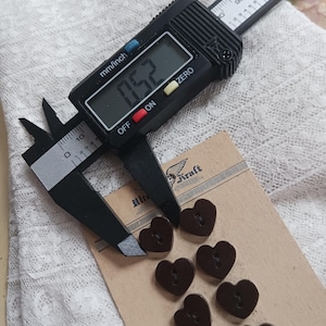 May include: A digital caliper measures a dark brown heart-shaped button. The button is approximately 0.52 inches in diameter. Several other heart-shaped buttons are visible on a Kraft paper card with a vintage logo.
