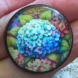May include: A round, floral-themed cabochon with a black metal bezel. The design features a vibrant hydrangea in shades of blue, pink, and white, surrounded by green leaves. The image is detailed and appears to be under a glass or resin dome.