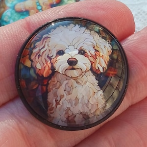 May include: A circular cabochon button featuring a detailed illustration of a fluffy, cream-colored dog with curly fur, dark eyes, and a dark nose, set within a smooth, black frame that encircles the artwork.
