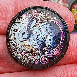 May include: A round, black-rimmed cabochon with a stained-glass style design. The image features a white rabbit with blue shading, surrounded by floral elements and a star. The design is set against a light background with a dark border.