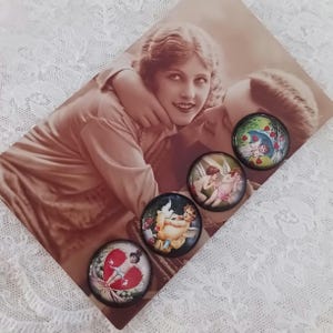 May include: Four round, black-rimmed magnets with vintage angel and heart designs are displayed on a sepia-toned photograph of a couple. The magnets feature cherubs, hearts, and floral elements in a romantic, antique style. The background is a white lace fabric.