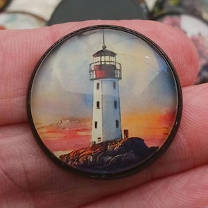 May include: A round, black-rimmed pendant featuring a watercolor painting of a white lighthouse with a red top. The lighthouse stands on a rocky outcrop, set against a sunset sky with orange and pink hues.