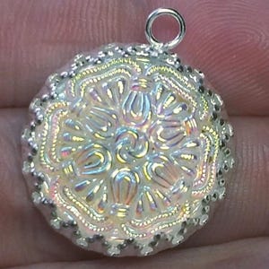 May include: A round pendant featuring an iridescent, milky white surface with a raised, symmetrical floral pattern. The circular charm is bordered by a textured, silver-toned metallic edge and includes a small, silver-toned jump ring at the top.