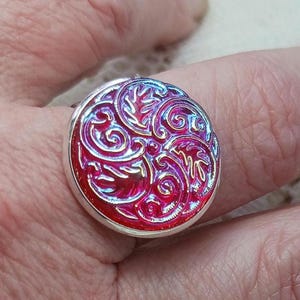 May include: A silver ring with a round, iridescent red and blue glass cabochon featuring a swirling, embossed floral design.