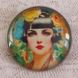 May include: A round, silver-toned brooch featuring a vintage illustration of a woman with blue eyes, red lips, and floral accents in her hair. The woman's portrait is set against a backdrop of green and yellow foliage.
