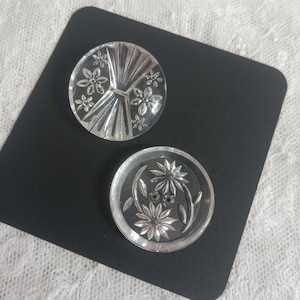 May include: Two clear, round buttons with floral designs. One button features a fan-like pattern with small flowers, while the other has a floral and leaf design. The buttons are on a black surface.