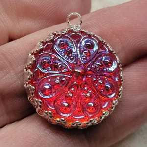 May include: A round, red pendant with a silver-colored border and a loop at the top. The pendant has a detailed, textured design with a floral pattern. The red color has an iridescent quality, with hints of purple and blue.